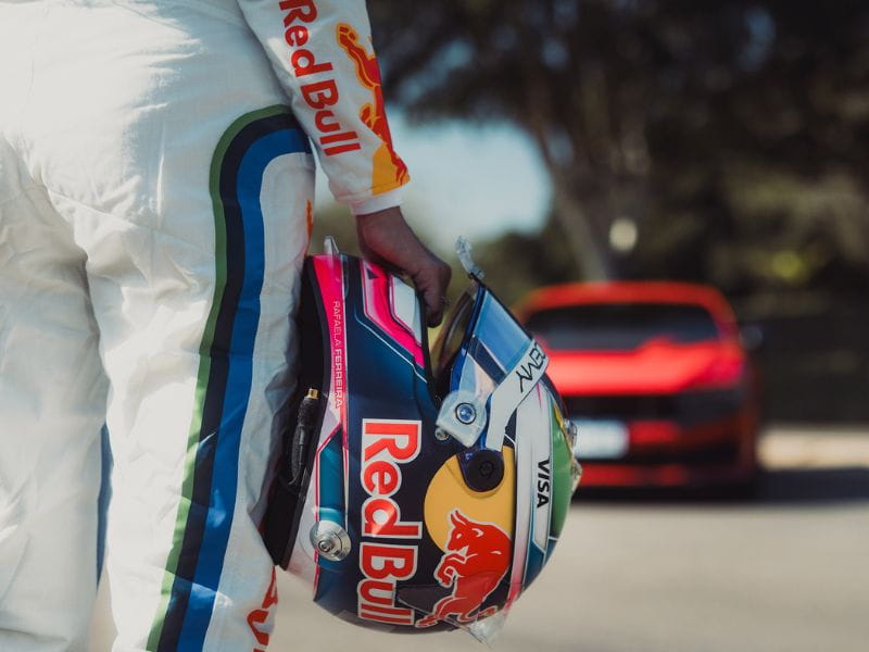 Uniforme e capacete de corrida com patrocínio Red Bull em frente a Ford Mustang Dark Horse