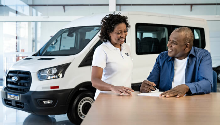 Homem e Mulher Sorrindo e Assinando um Contrato, com Transit Minibus ao Fundo, representando Ford Pro Financiamento