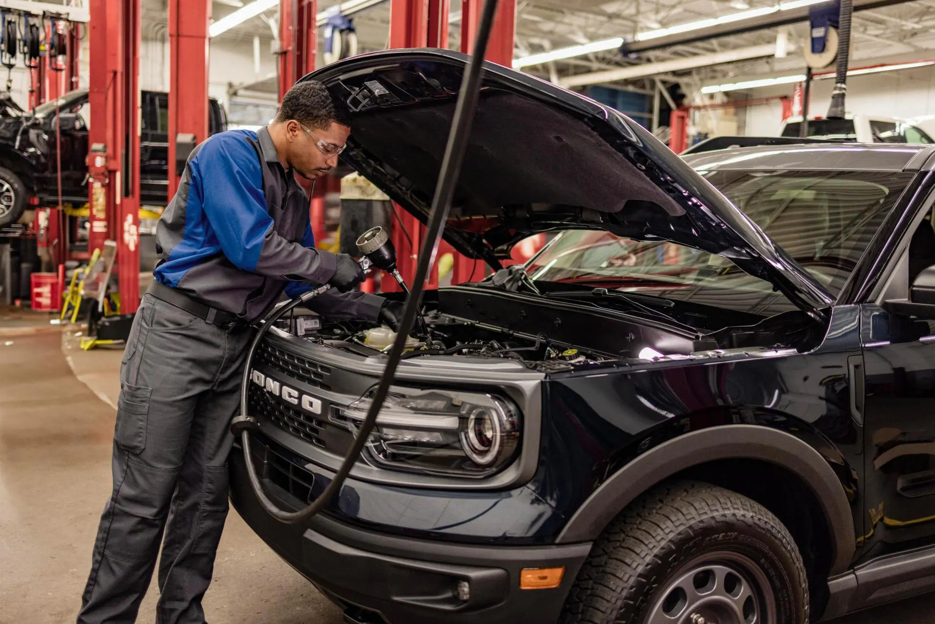 Especialista da Ford Dimas realizando um procedimento em um veículo com o capô aberto. Ele está trabalhando no motor do carro e o ambiente é a oficina da assistência técnica da concessionária.