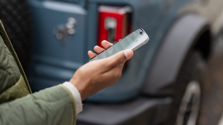 Una persona sosteniendo un celular y de fondo una Bronco azul