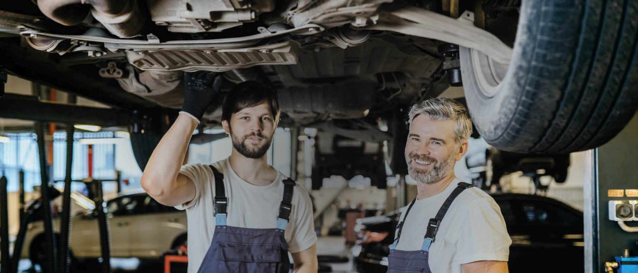 Karriere Garage Rütter Zwei Mechaniker in Garage stehen unter Carosserie eines Fahrzeugs