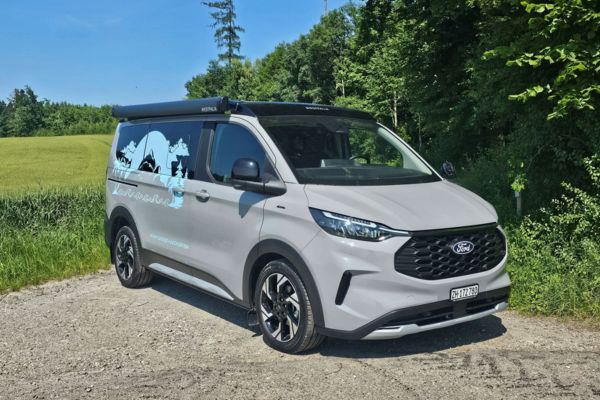 Grauer Transit Custom Nugget steht auf Kieselsteinen mit Wiese und Bäumen im Hintergrund