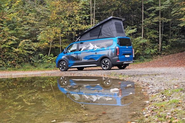 Blauer Transit Custom Nugget steht im Wald am Rande eines Sees der das Fahrzeug reflektiert