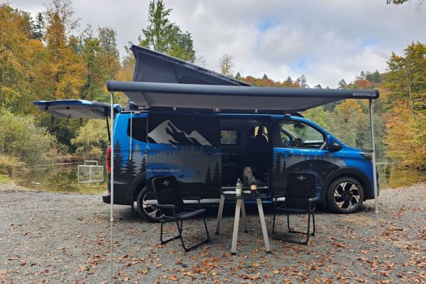 Blauer Transit Custom Nugget Plus mit Vordach steht auf Kieselsteinen