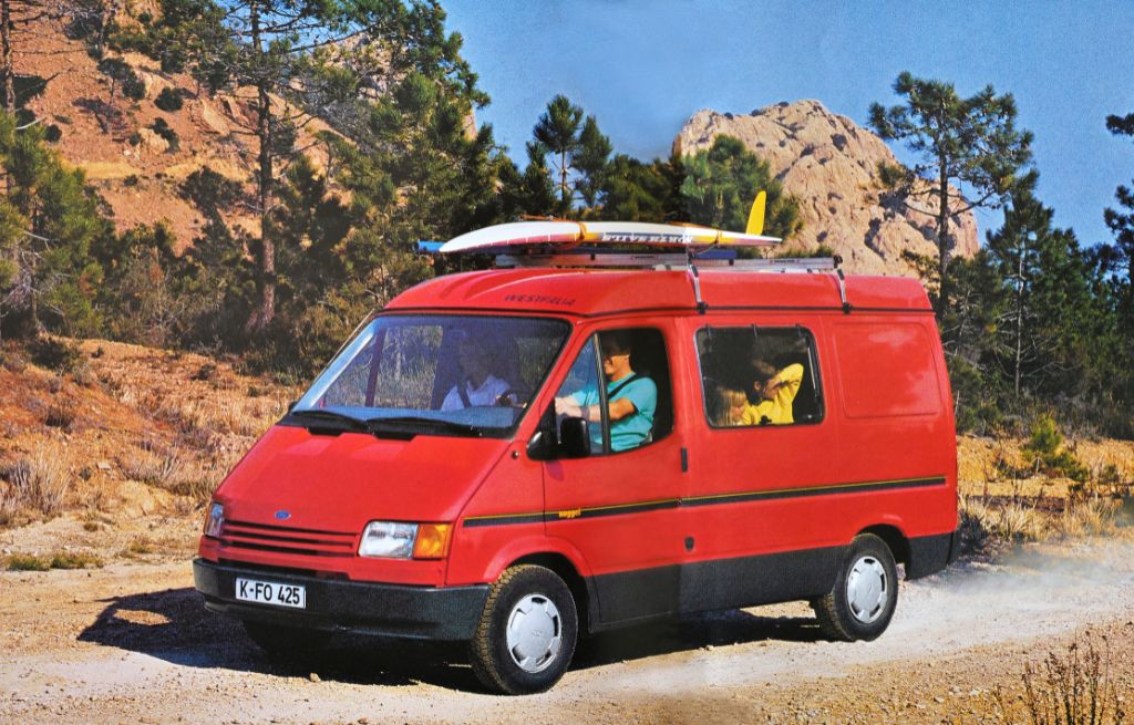 Ford Nugget von 1993 mit Dachträger und Surfbrett fährt auf staubiger Bergstrasse durch die Berge.