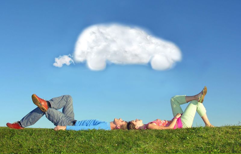 Couple lying on grass looking at car-shaped cloud in blue sky