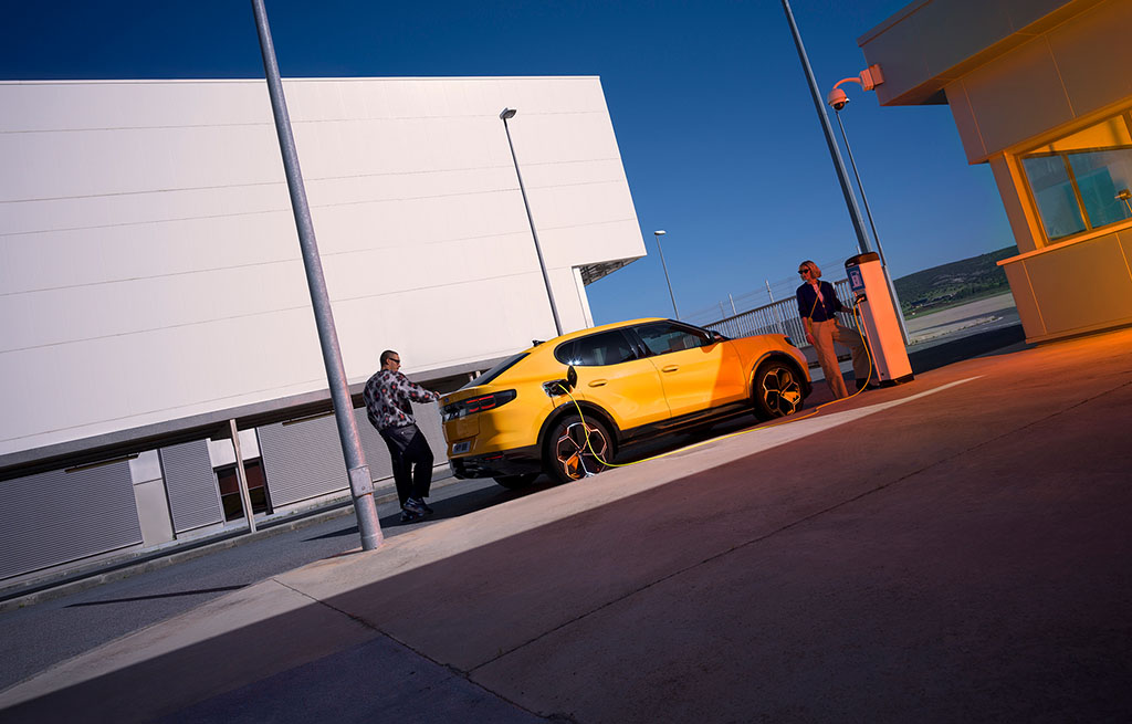 A yellow All-Electric Capri is parked at a public charging station with two people nearby, under a clear evening sky