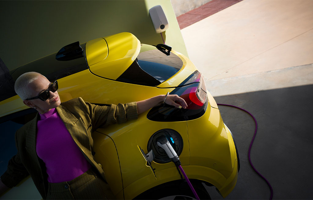 A woman wearing sunglasses and a purple top leans on the rear of a yellow electric car while plugging in a charging cable at a home charging station