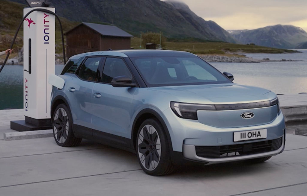 A light blue All-Electric Explorer SUV is parked at an IONITY charging station near a lake with mountains in the background, ready for charging
